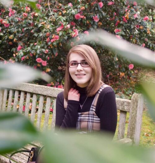 A photo of Elin sitting on a bench, she is framed by some green leaves which are out of focus