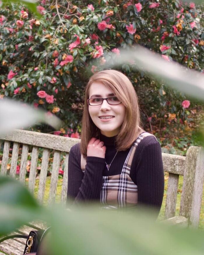 A photo of Elin sitting on a bench, she is framed by some green leaves which are out of focus