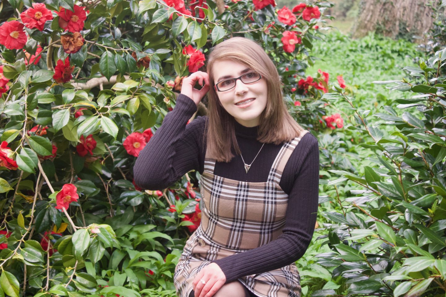 A photo of Elin crouching down in front of some red flowers on a green bush