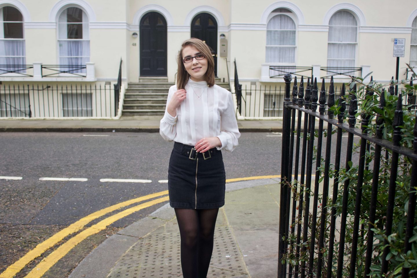 A portrait photo of Elin wearing a black denim double buckle skirt, a white high neck pleated blouse