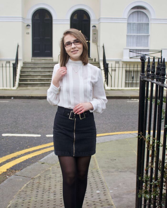 A portrait photo of Elin wearing a black denim double buckle skirt, a white high neck pleated blouse