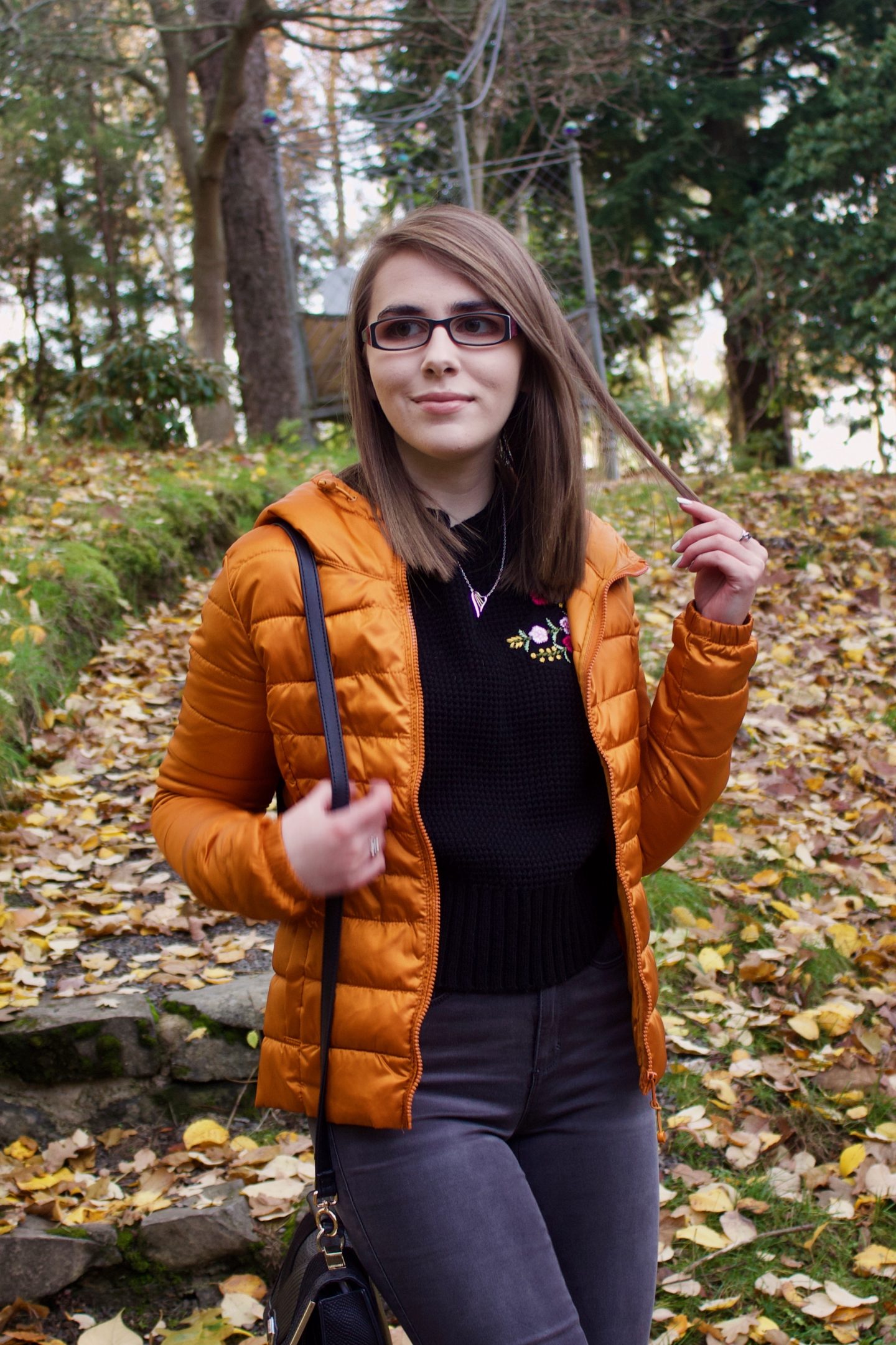 A photo of Elin wearing a black knitted jumper and orange/marmelade coloured quilted jacket, she is standing on some steps in a woodland and there are colourful leaves on the floor around her, her hand is up, holding a strand of hair and she is looking away from the camera