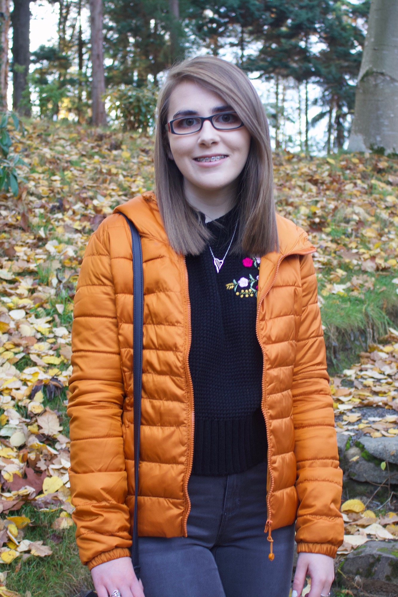 A photo of Elin wearing a black knitted jumper and orange/marmelade coloured quilted jacket, she is standing on some steps in a woodland and there are colourful leaves on the floor around her.