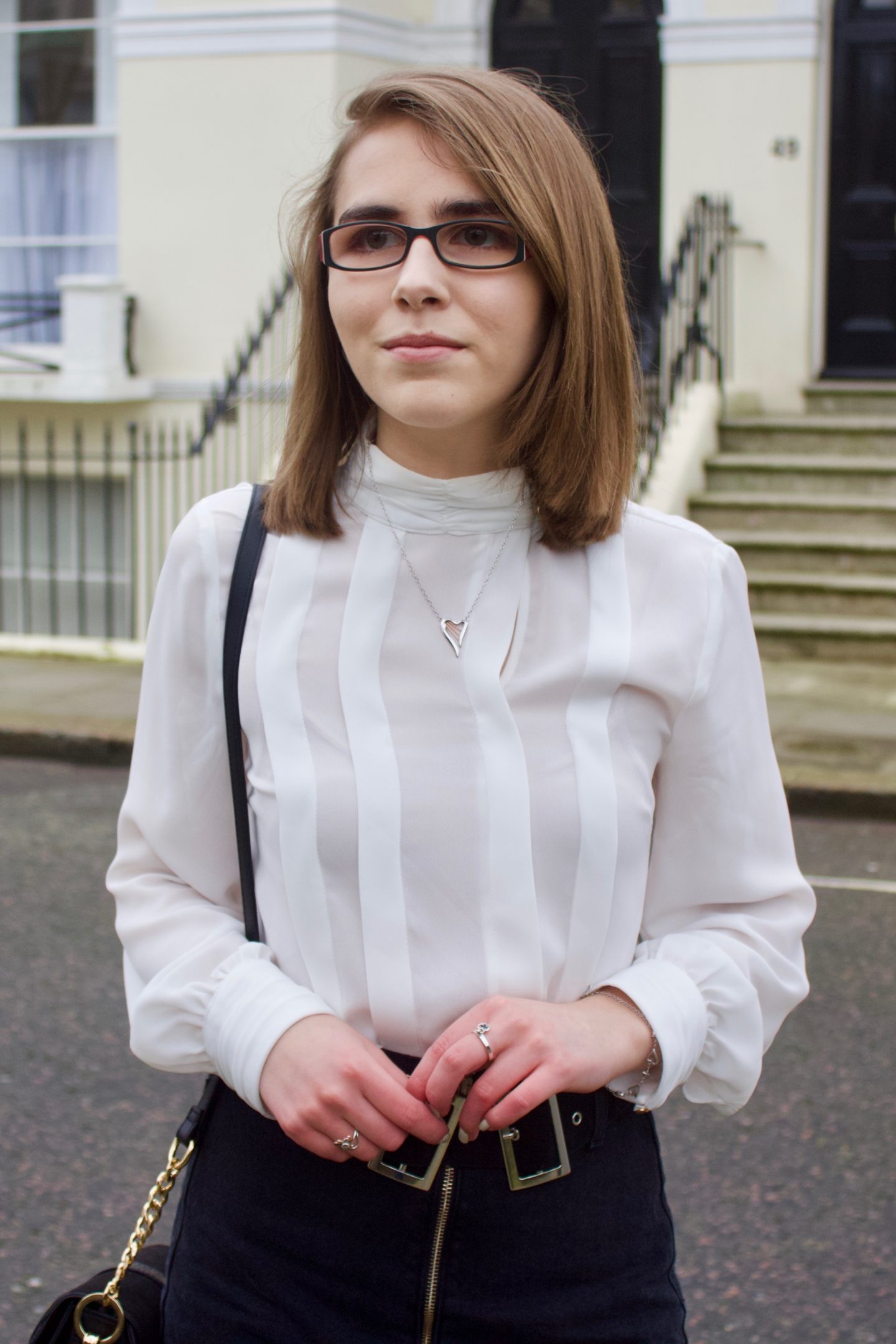 A photo of Elin looking away from the camera, she is wearing a white pleated high neck blouse and is standing in front of some fancy London houses which have steps leading up to them
