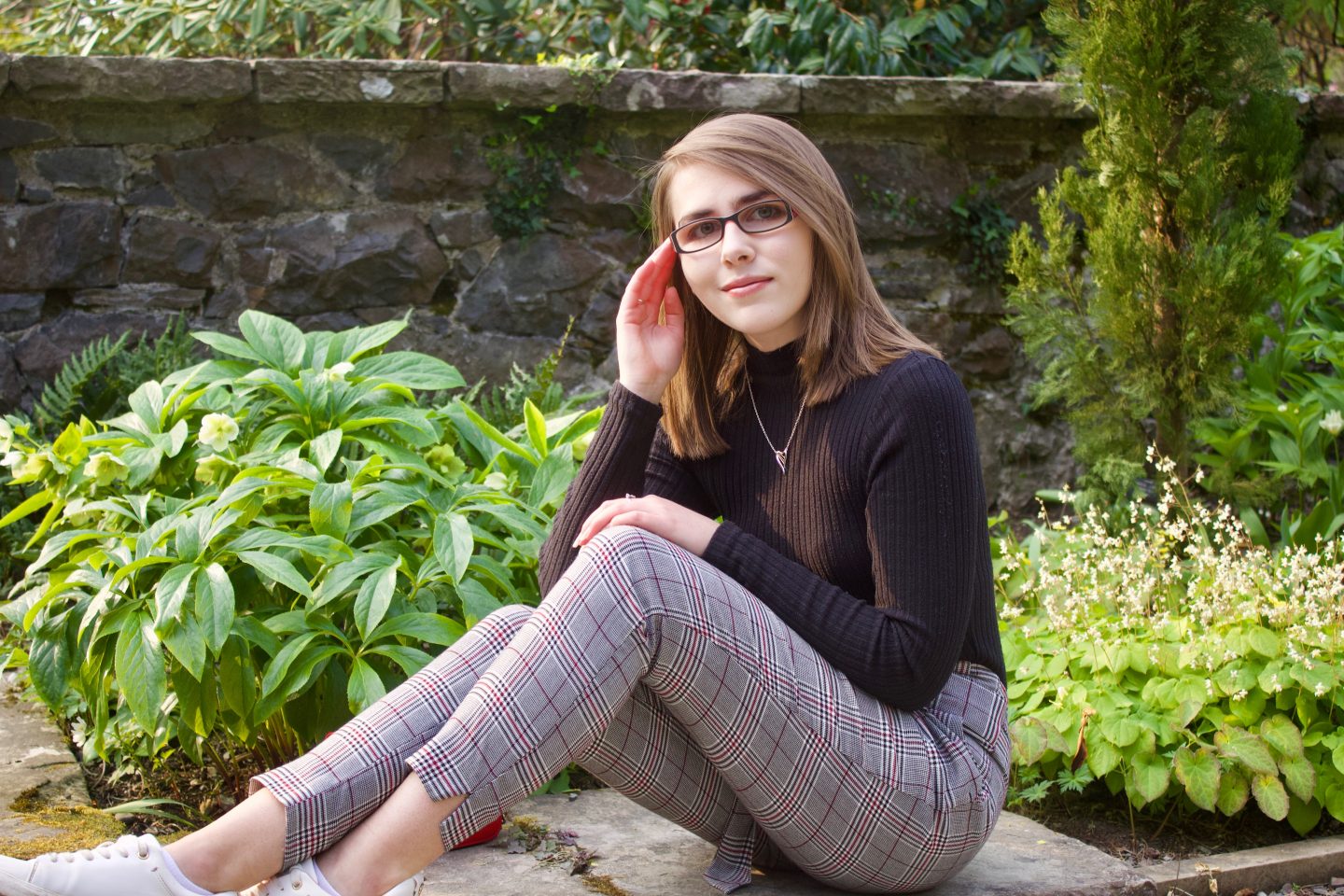 Elin wearing grey check trousers and a black high neck top, there are green plants behind her. She's sitting on a stone wall, her left hand rests on her left knee whilst her right elbow rests on her right knee and her hand is raised to rest on the arm of her glasses.
