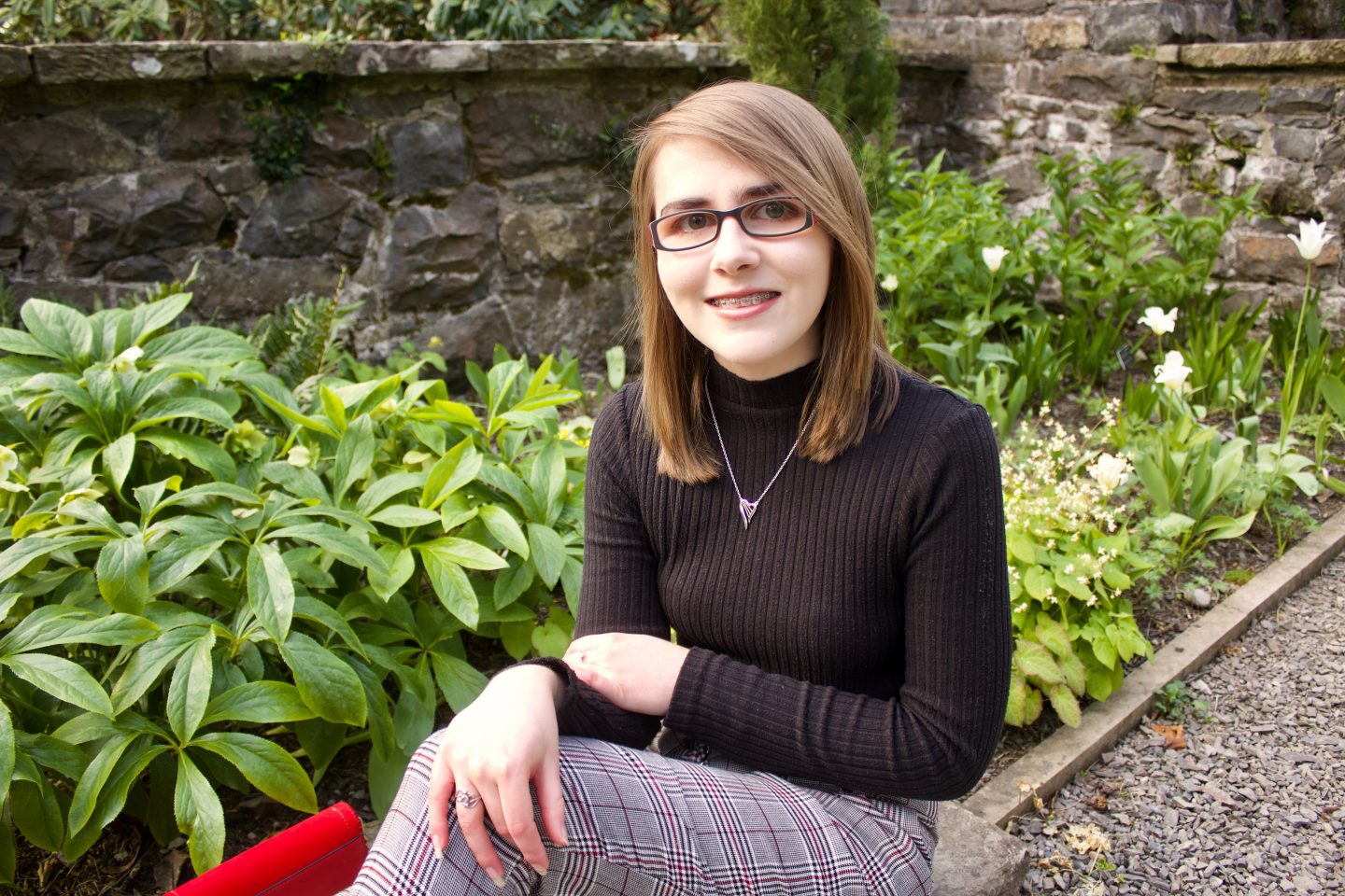 A photo of Elin sitting in front of green plants with a stone wall in the background