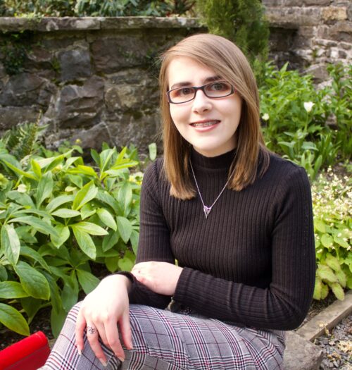 A photo of Elin sitting in front of green plants with a stone wall in the background