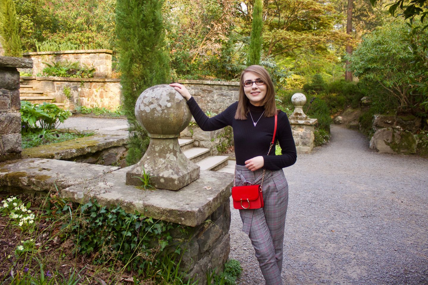 Elin wearing check skinny trousers, a black high neck top and small red handbag with a gold ring on the front. She rests her hand on a stone wall beside her and she's smiling at the camera.