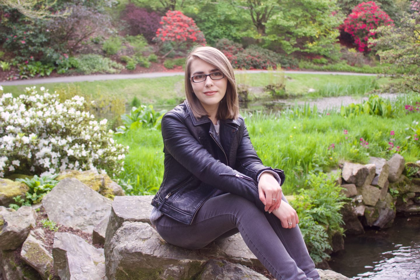 A photo of Elin wearing a grey knitted jumper, black leather jacket and grey jeans, she is sitting on a rock and there are colourful flowers in the background