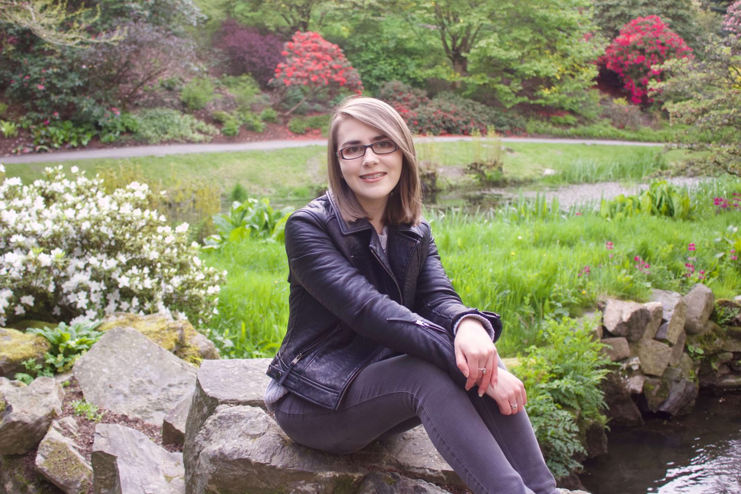 A photo of Elin wearing a grey knitted jumper, black leather jacket and grey jeans, she is sitting on a rock and there are colourful flowers in the background
