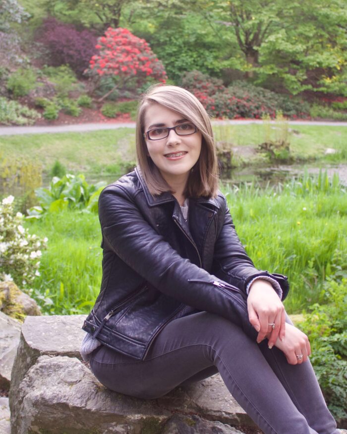 A photo of Elin wearing a grey knitted jumper, black leather jacket and grey jeans, she is sitting on a rock and there are colourful flowers in the background