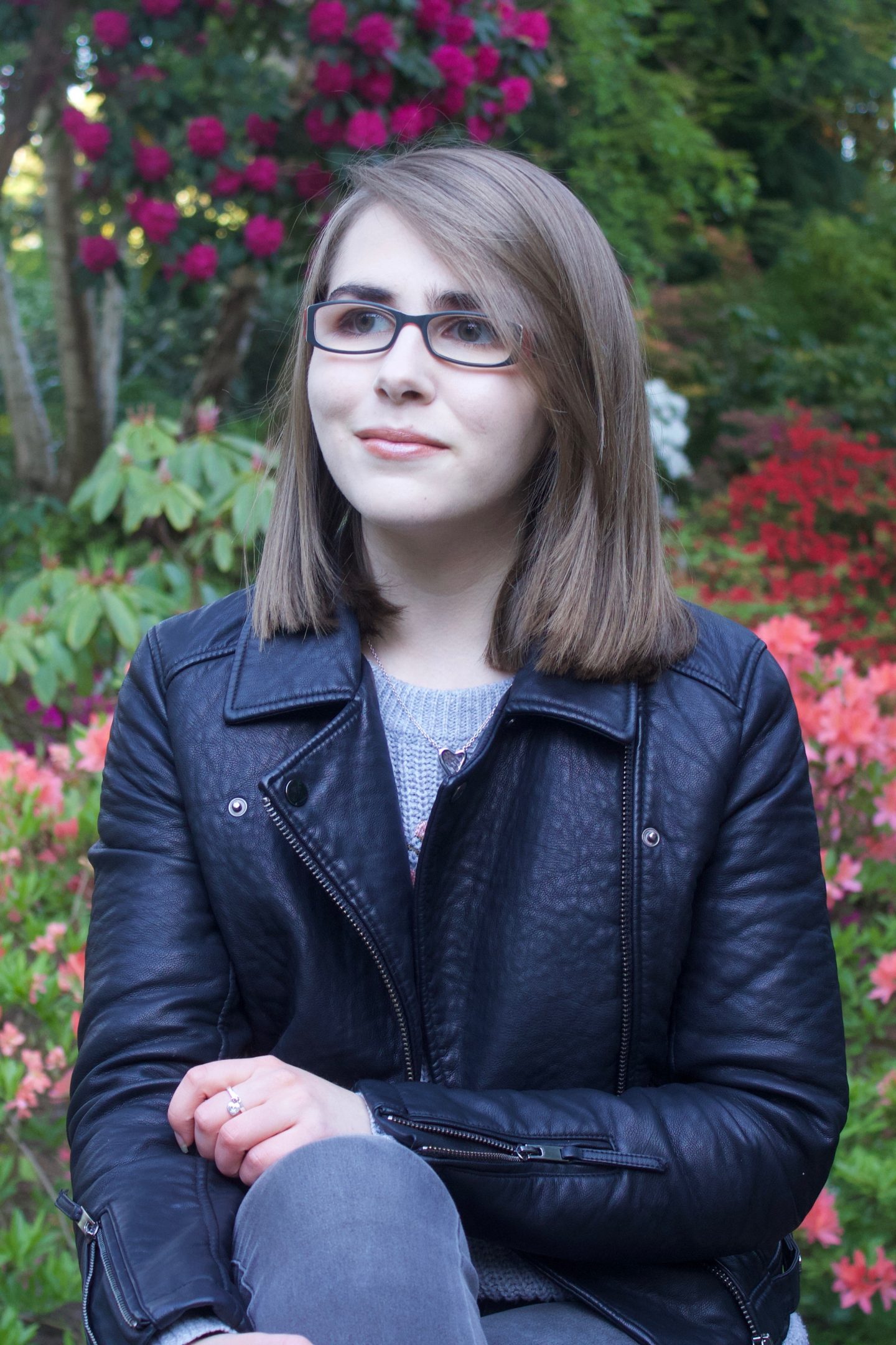A photo of Elin wearing a grey knitted jumper and black faux leather jacket, she is sitting on some rocks with colourful flowers in the background