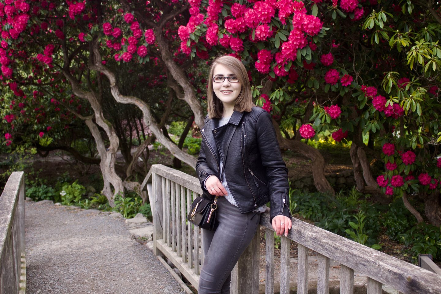 A photo of Elin leaning against a wooden bridge and smiling at the camera. There are pink flowers on trees in the background