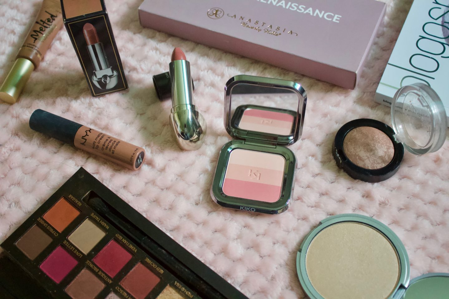 A photo of miscellaneous beauty products including a pink eyeshadow palette, a highlighter and a lipstick