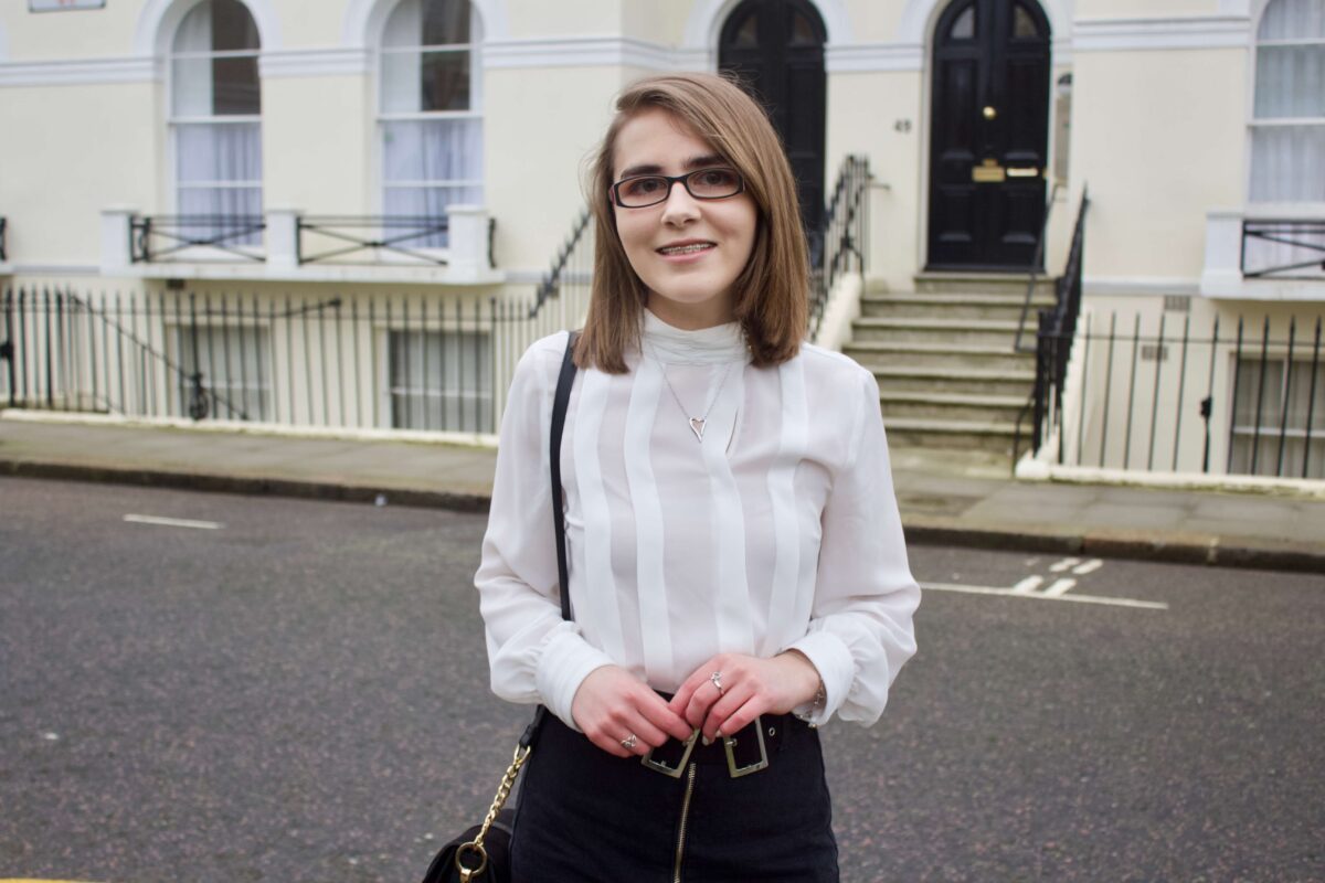 A photo of Elin wearing a white high neck pleated blouse, a zip up denim belt which has a double buckle belt and a small black cross body bag