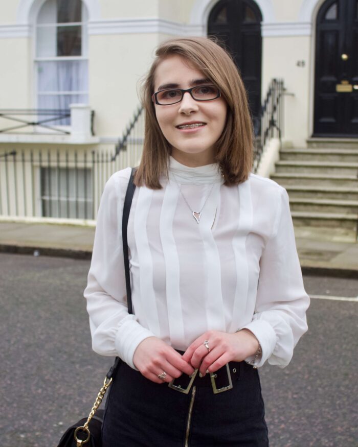 A photo of Elin wearing a white high neck pleated blouse, a zip up denim belt which has a double buckle belt and a small black cross body bag