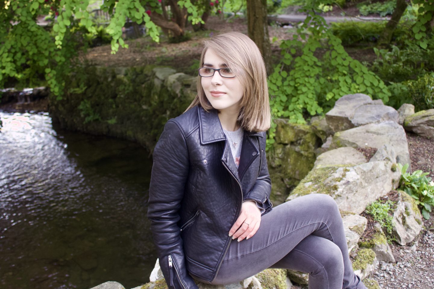 A photo of Elin sitting on a small stone wall with water and trees in the background