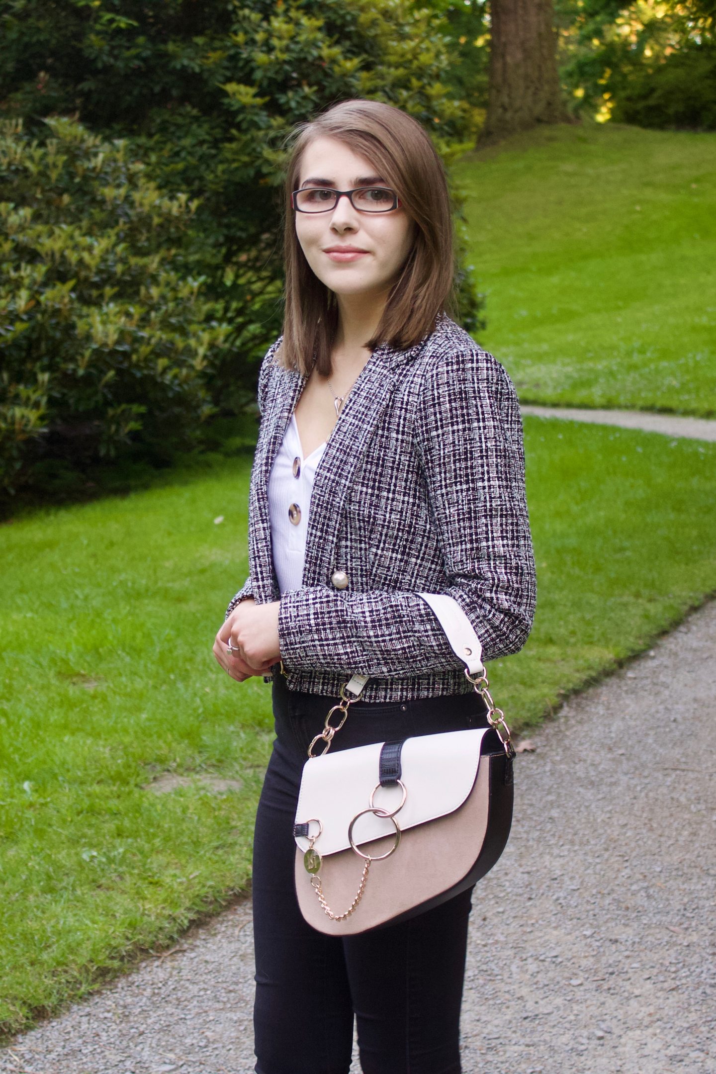 A photo of Elin from the side, she wear a grey boucle jacket with a white v-neck top which has buttons on the neckline, black skinny jeans and a white and beige saddle bag