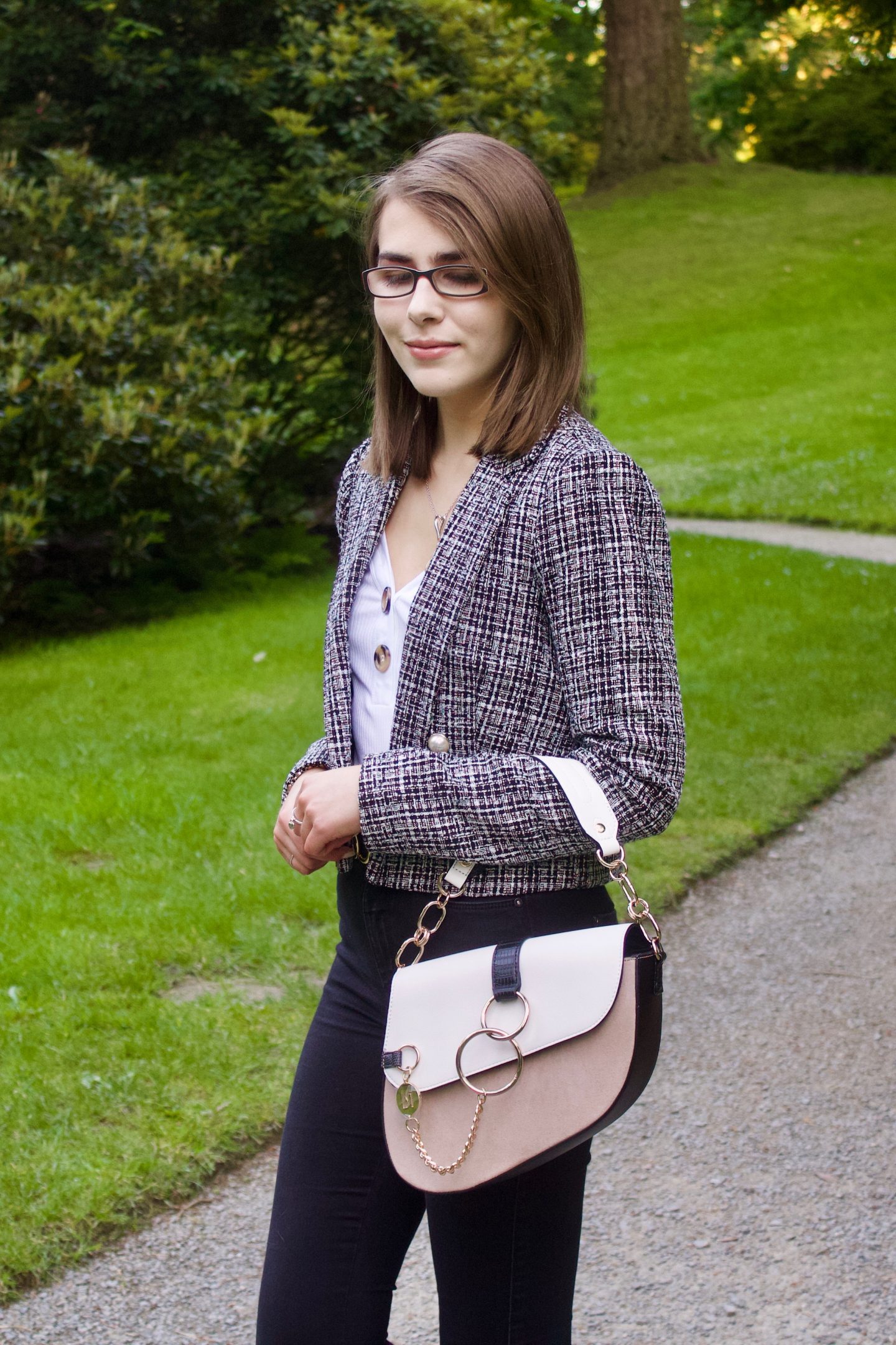 A photo of Elin from the side, she wear a grey boucle jacket with a white v-neck top which has buttons on the neckline, black skinny jeans and a white and beige saddle bag, she is looking down