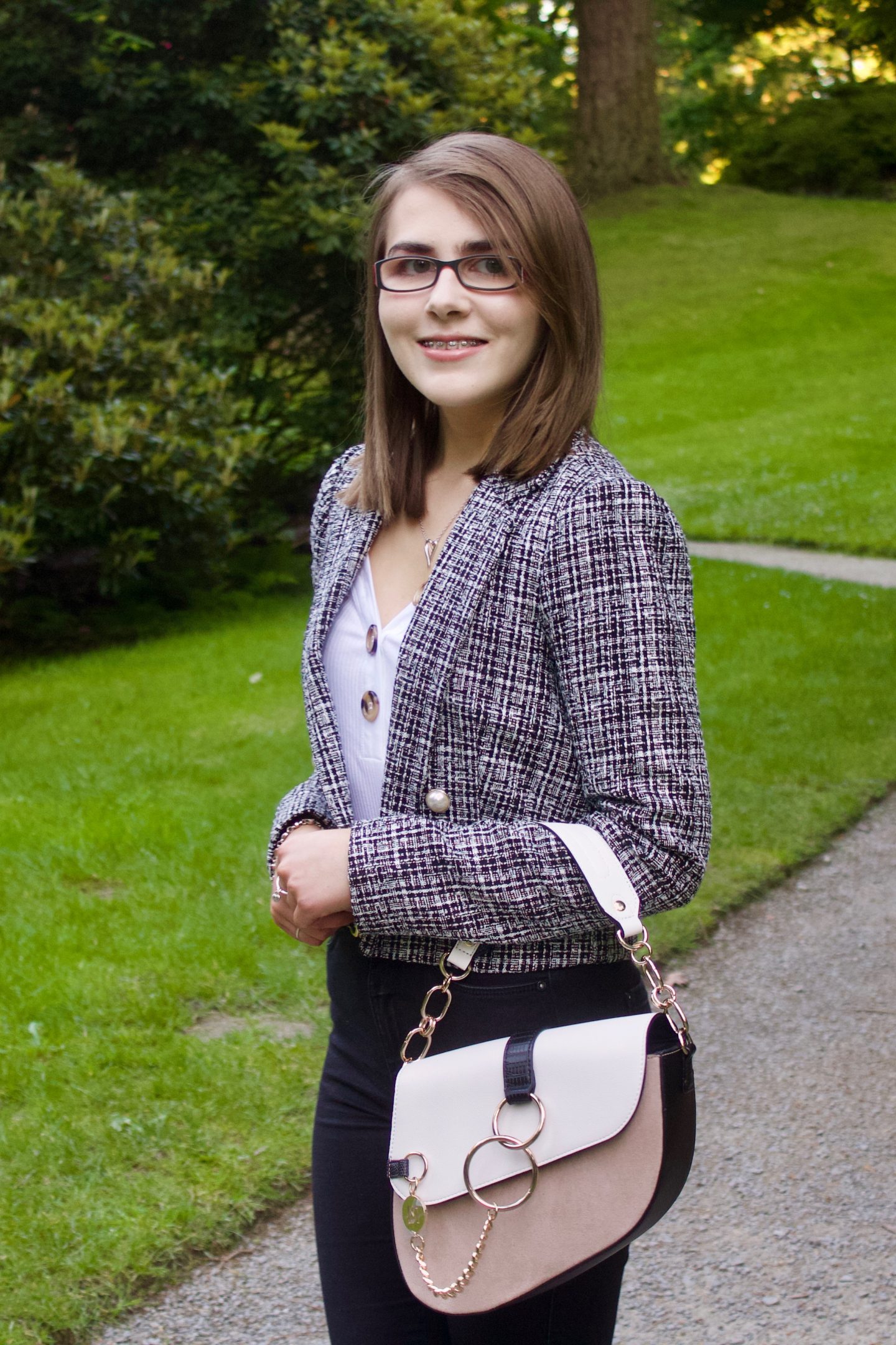 A photo of Elin from the side, she wear a grey boucle jacket with a white v-neck top which has buttons on the neckline, black skinny jeans and a white and beige saddle bag, she is smiling at the camera