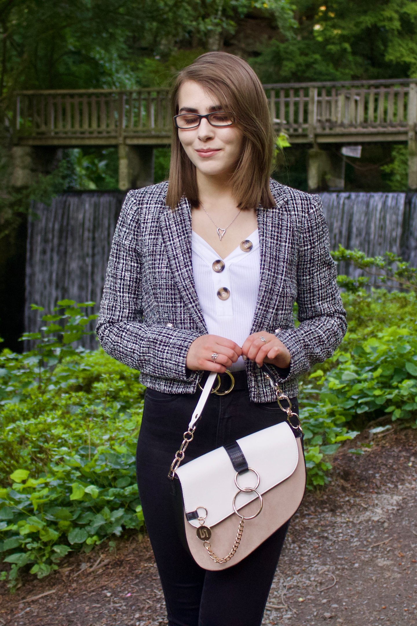 A photo of Elin looking down, she wears a grey boucle cropped jacket, a white v-neck top, black skinny jeans and a white and beige saddle bag. A waterfall can be seen in the background.