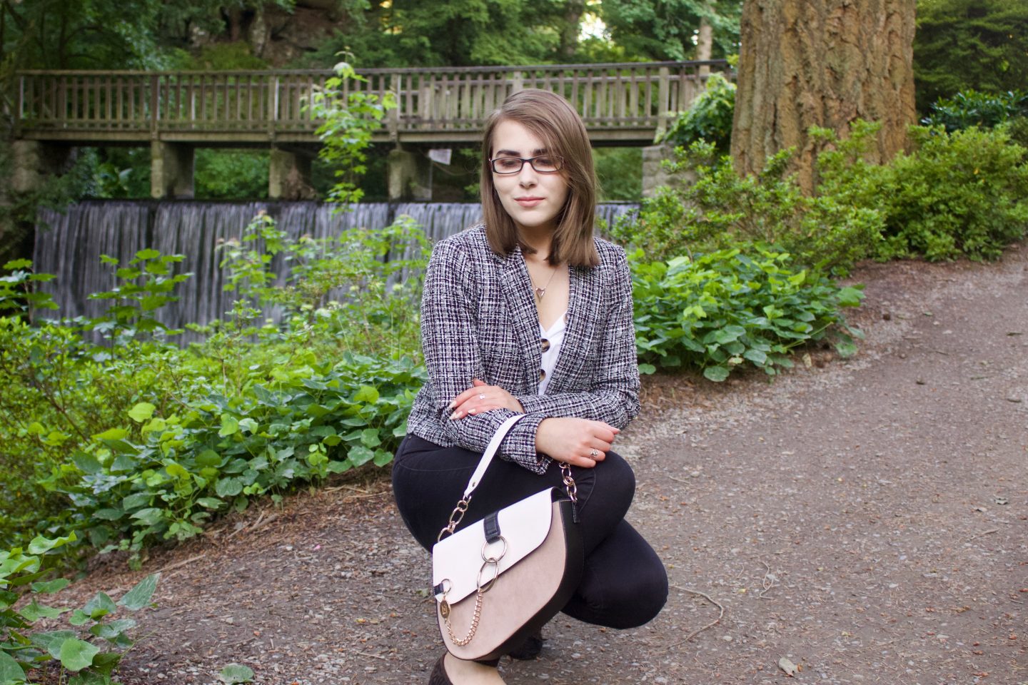 A photo of Elin crouching down and also looking down, there are flowers in the background as well as a wooden bridge with a waterfall underneath