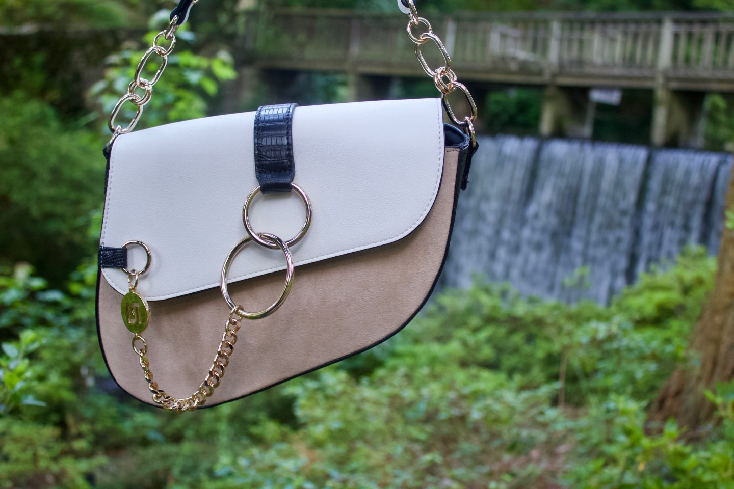 A photo of a white and beige saddle bag which has gold hardware, there is a waterfall in the background