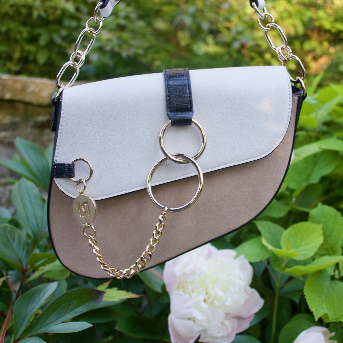 A photo of a white and beige saddle bag which has gold hardware, there are white flowers in the background