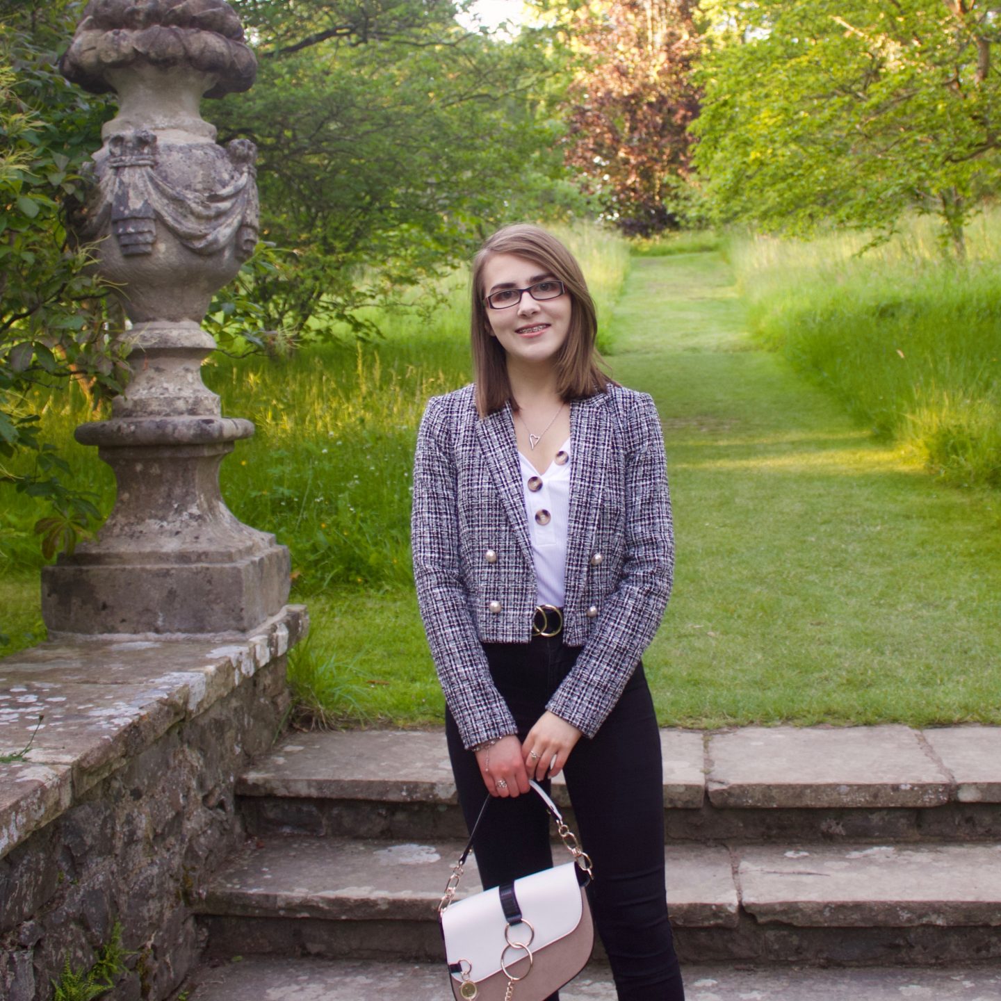 A photo of Elin standing on stone steps which lead to a green patch of land, she is wearing a white v-neck top which has buttons on the neckline, a grey boucle jacket, black skinny jeans and she's holding a white and beige saddle back which has gold hardware
