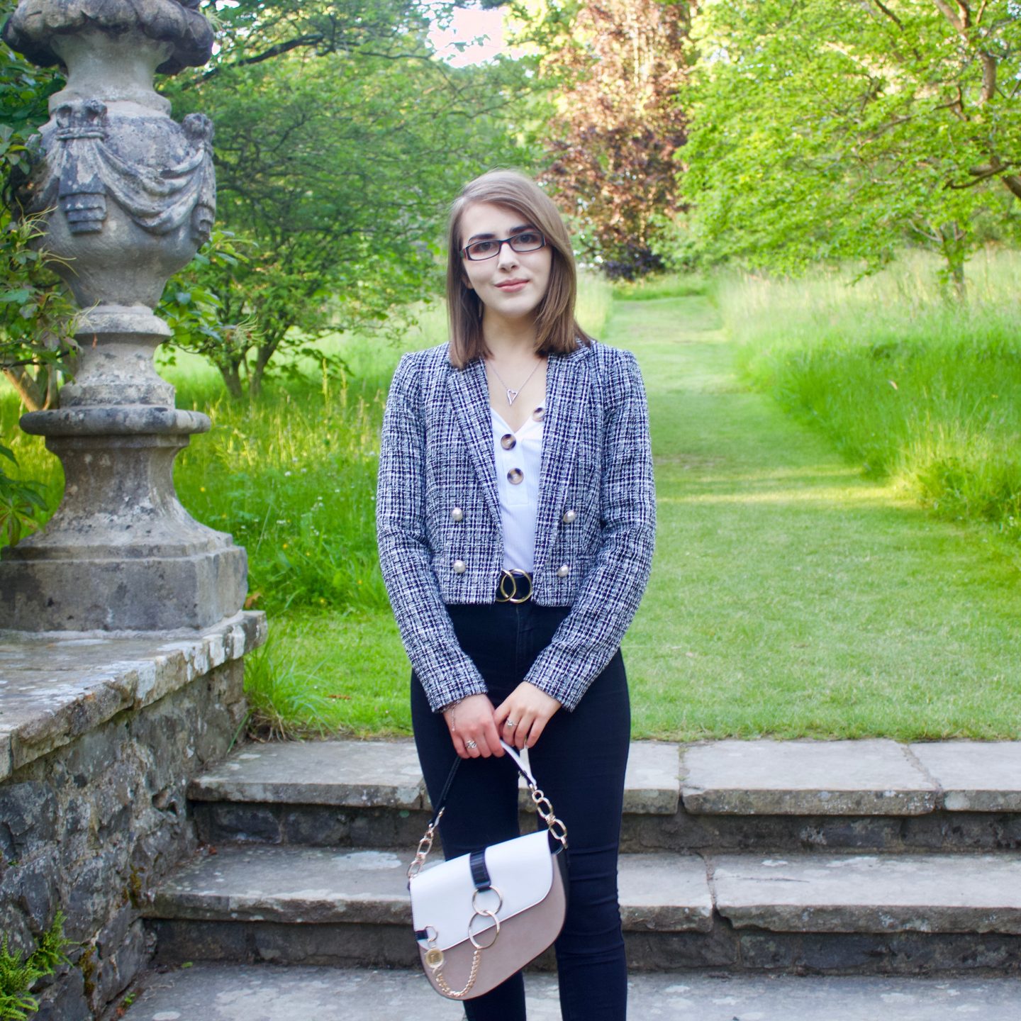 A photo of Elin standing on stone steps which lead to a green patch of land, she is wearing a white v-neck top which has buttons on the neckline, a grey boucle jacket, black skinny jeans and she's holding a white and beige saddle back which has gold hardware
