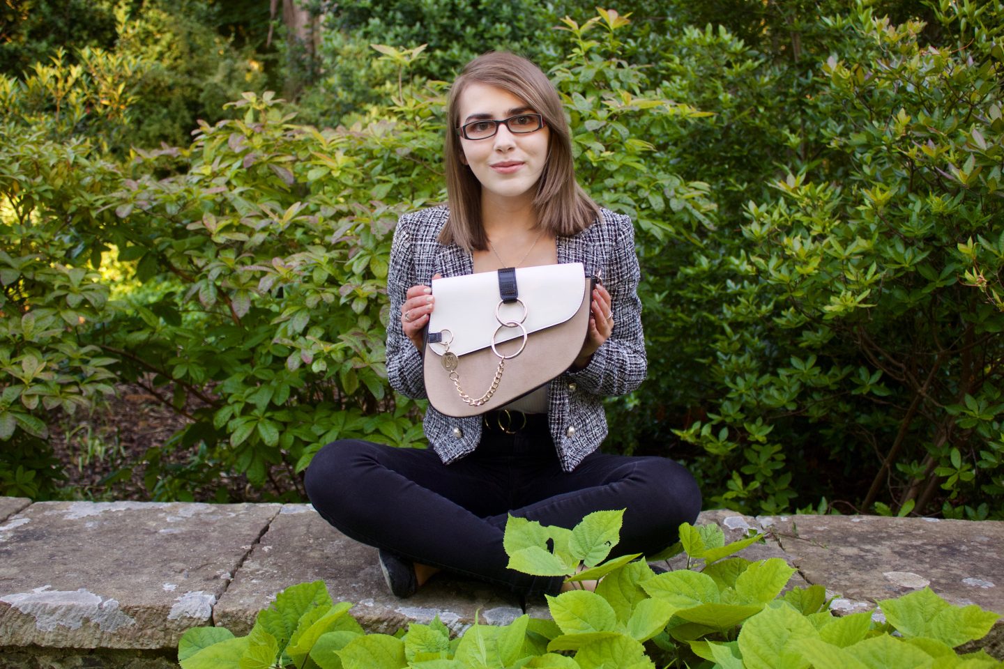 A photo of Elin sitting cross legged on a stone wall, she's holding a white and beige saddle bag in front of her