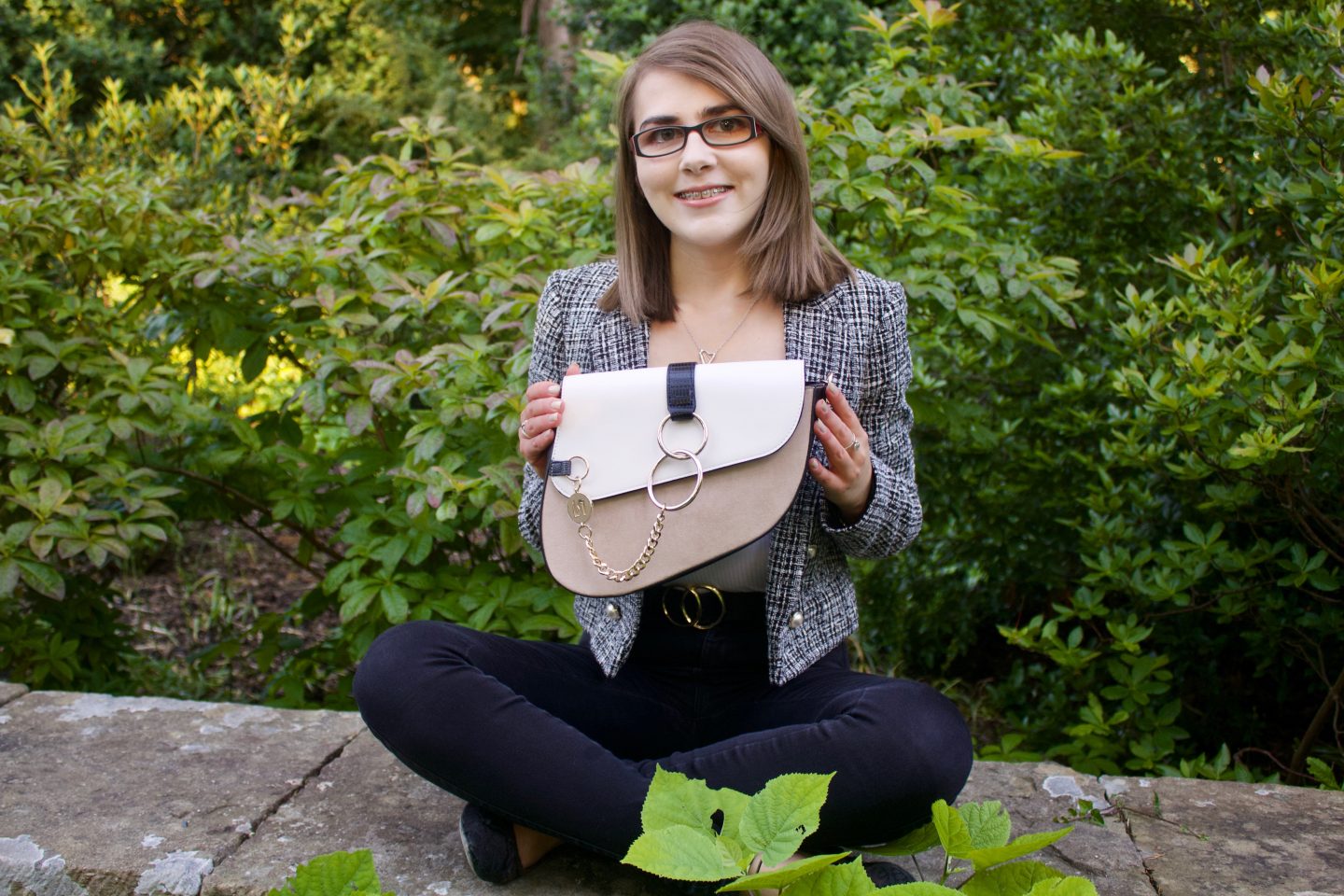 A photo of Elin sitting cross legged on a stone wall, she is smiling at the camera and holding a white and beige saddle bag in front of her