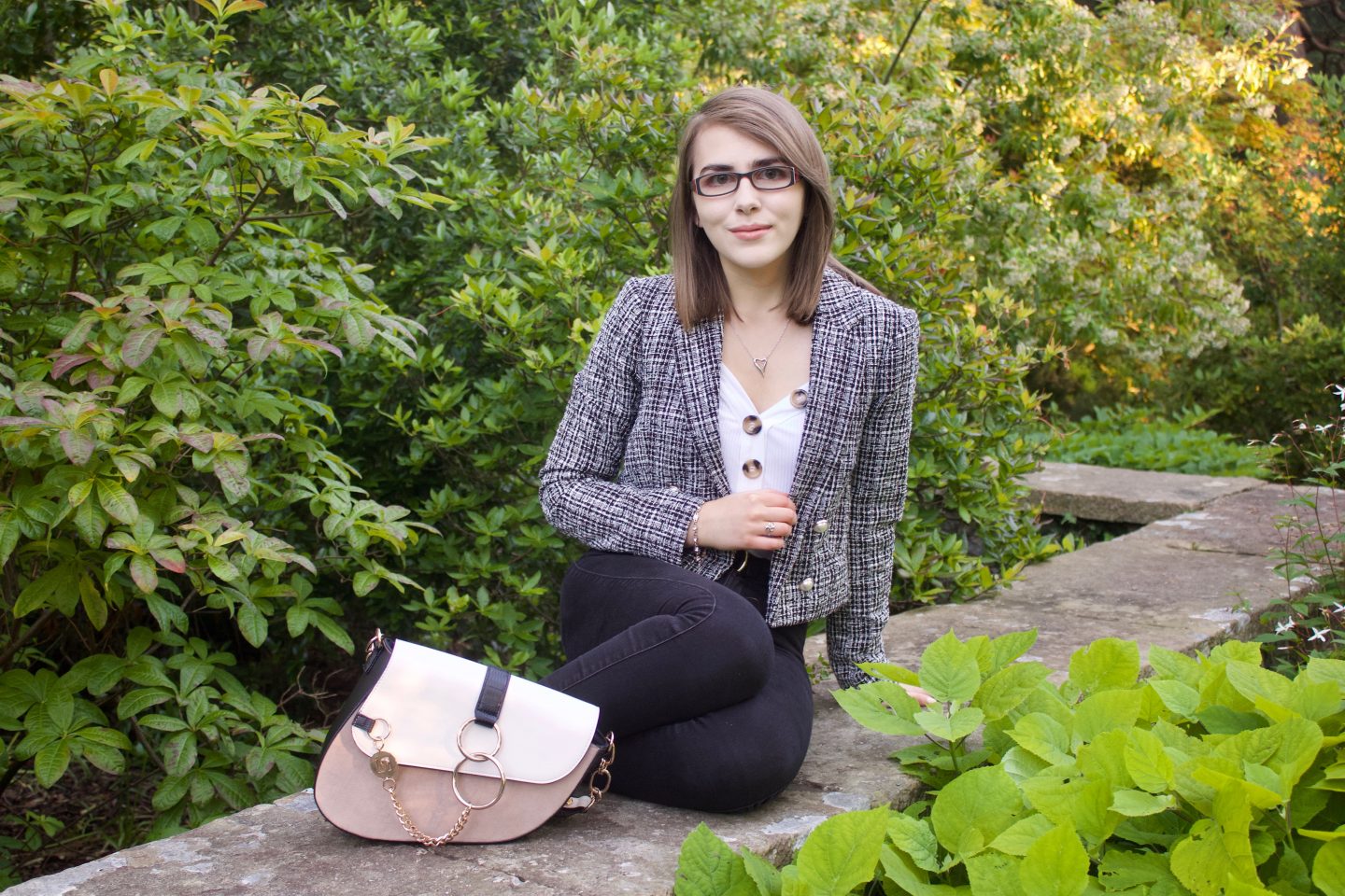A photo of Elin sitting on a stone wall with green trees and flowers in the background