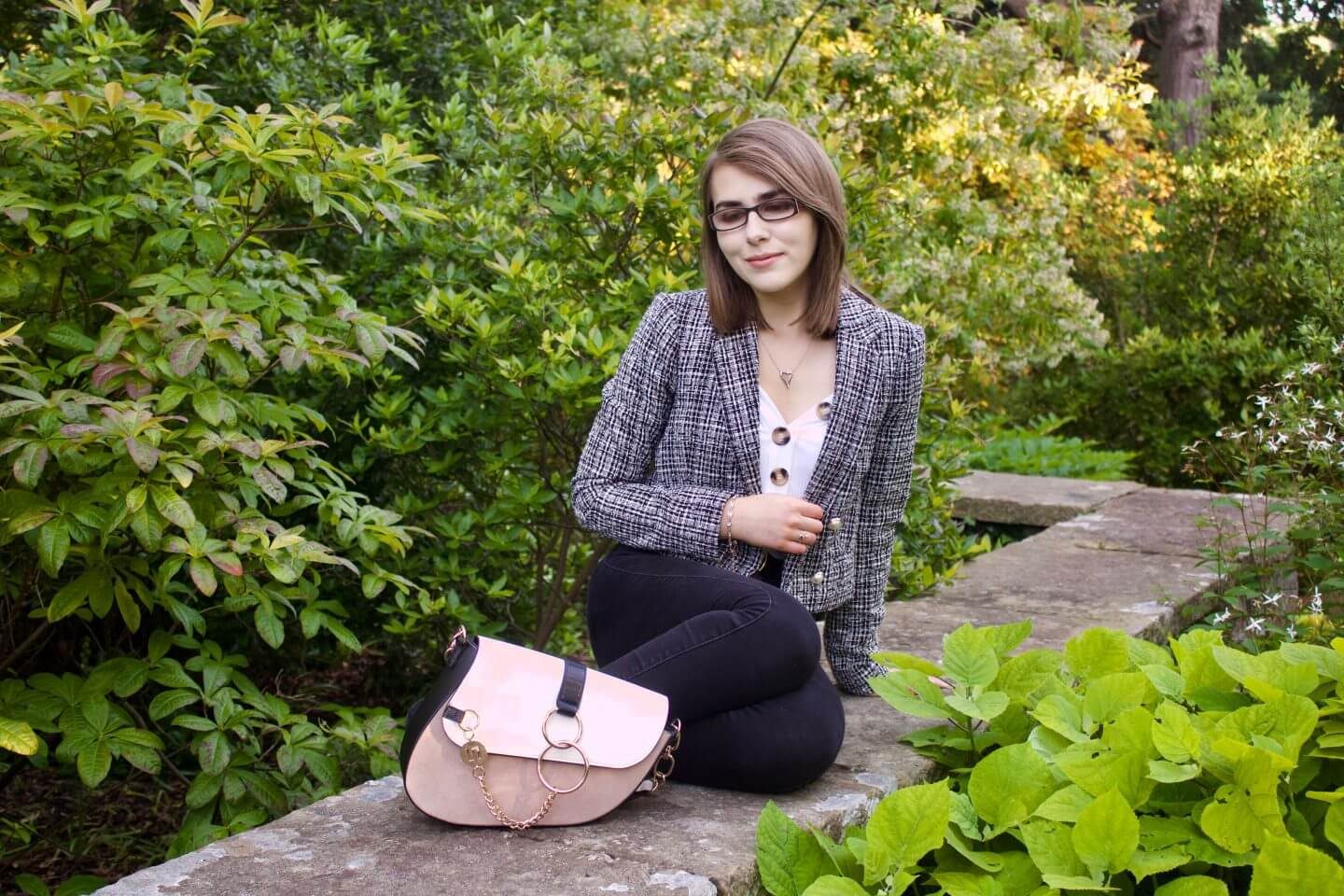 A photo of Elin sitting on a stone wall with green flowers in front of it and behind, she is wearing a grey boucle jacket, a white v neck top with gold buttons and black skinny jeans, her bag is placed in front of her and she is looking down
