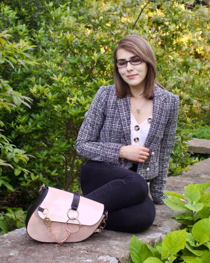 A photo of Elin sitting on a stone wall with green flowers in front of it and behind, she is wearing a grey boucle jacket, a white v neck top which has buttons on the neckline and black skinny jeans, her bag is placed in front of her and she is looking down