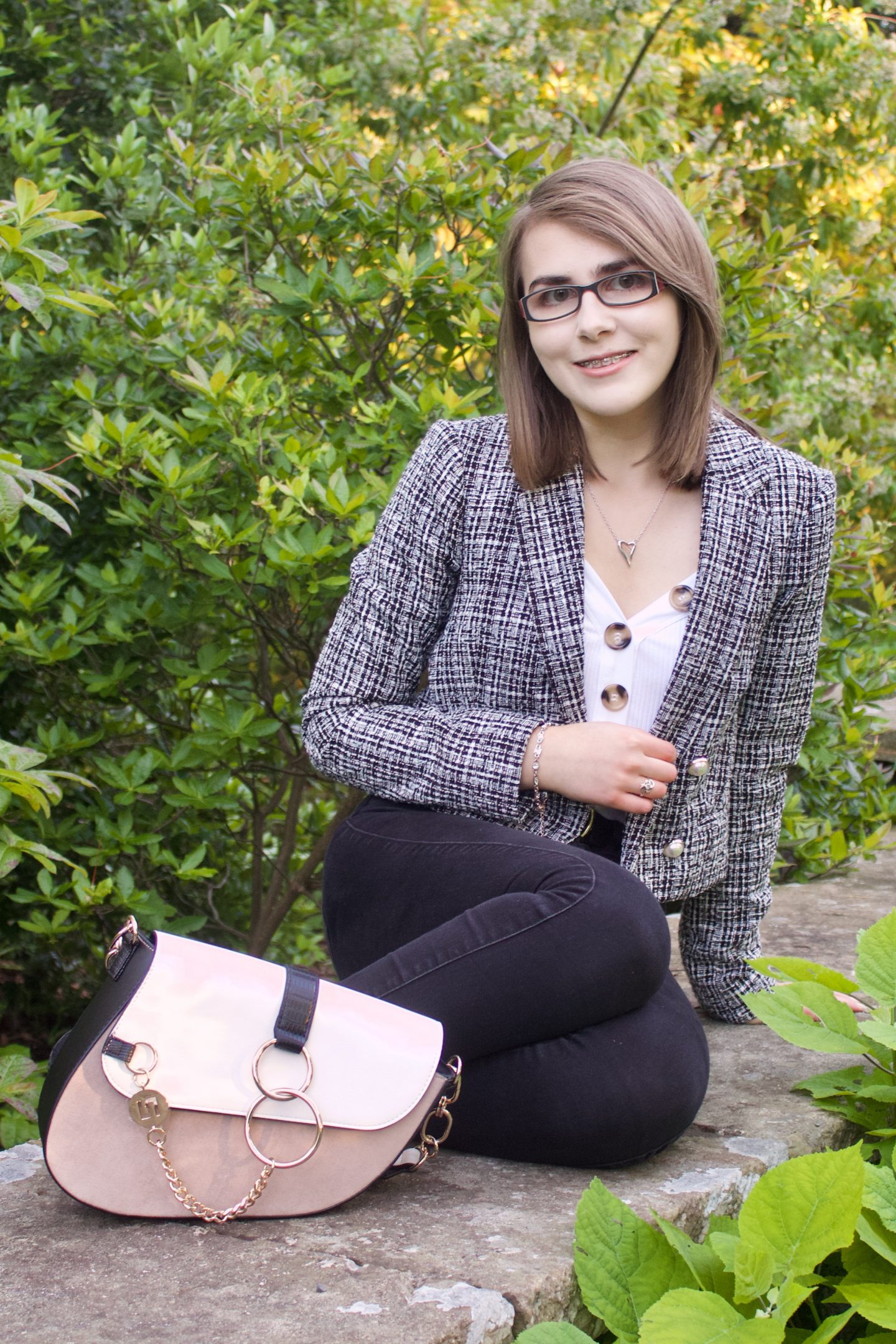 A photo of Elin sitting on a stone wall with green flowers in front of it and behind, she is weearing a grey boucle jacket, a white v neck top with gold buttons and black skinny jeans, her bag is placed in front of her and she is smiling at the camera