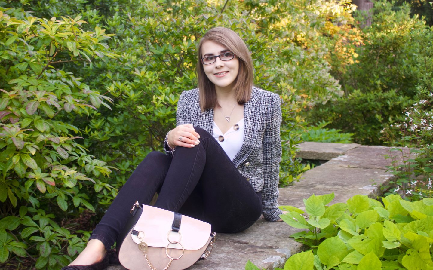 A photo of Elin sitting on a stone wall with green flowers in front of it and behind, she is wearing a grey boucle jacket, a white v neck top with gold buttons and black skinny jeans, her bag is placed in front of her and she is looking down