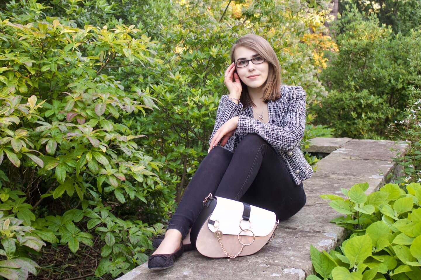 A photo of Elin sitting on a stone wall with green flowers in front of it and behind, she is wearing a grey boucle jacket, a white v neck top with gold buttons and black skinny jeans, her bag is placed in front of her