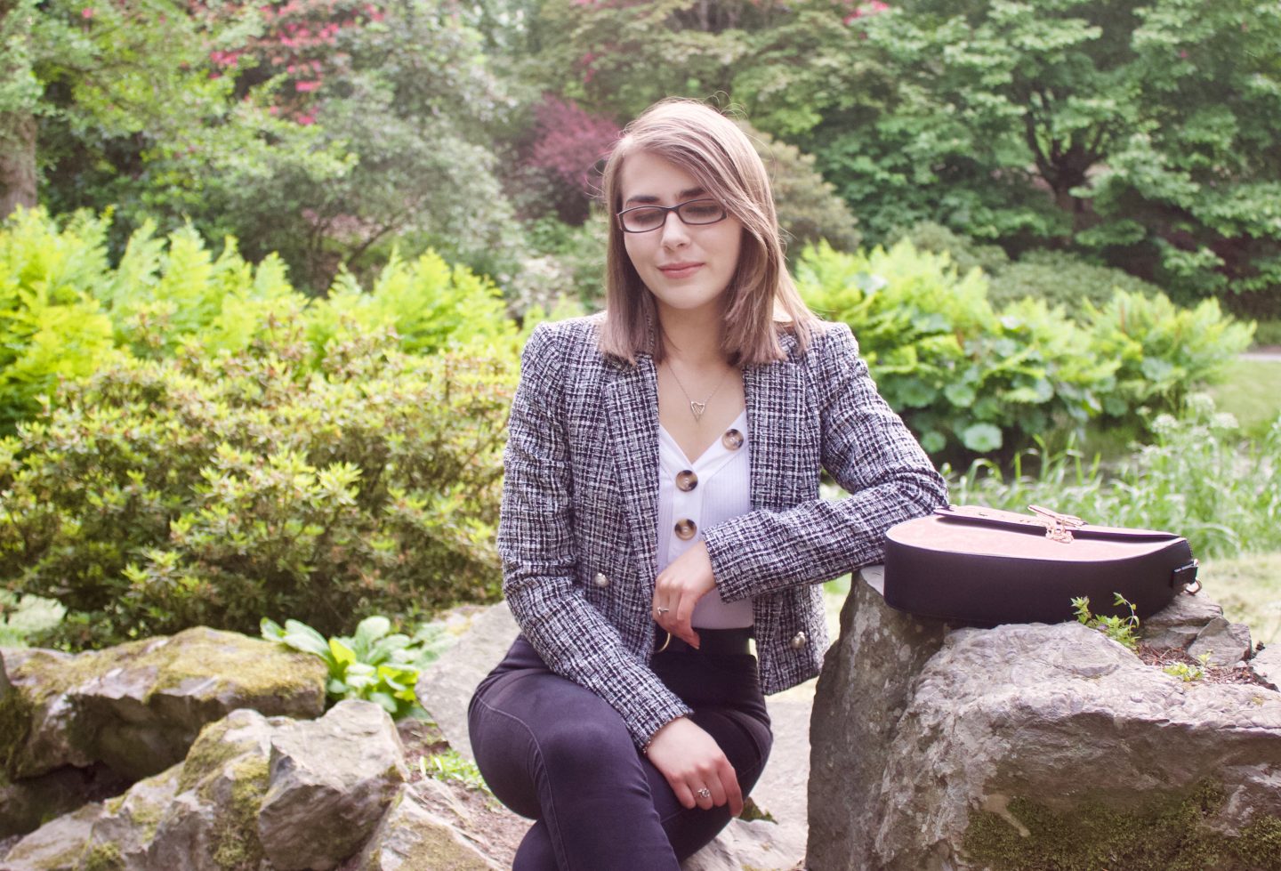 Disability & employment: A photo of Elin sitting on a stone whilst leaning against another, she is looking down and there are green plants and flowers in the background