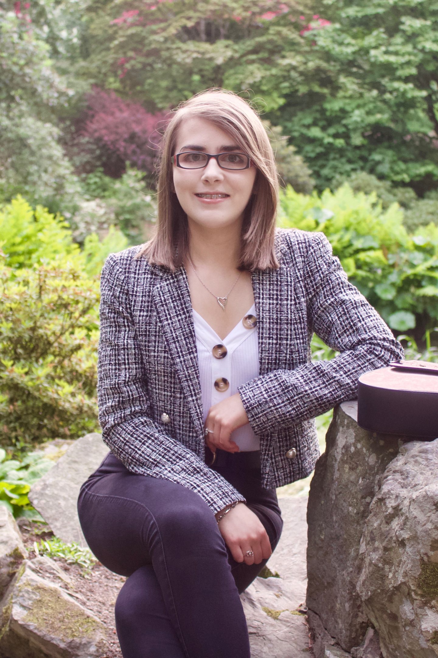 A photo of Elin sitting on a small rock whilst leaning against a bigger one which her bag is placed on, she is smiling at the camera