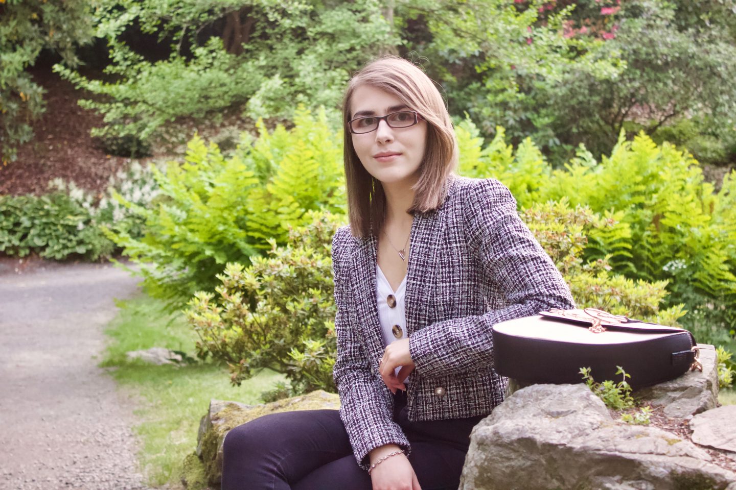 Disability & employment: A photo of Elin sitting on a rock with her arm leaning on another rock, there are green plants and flowers in the background