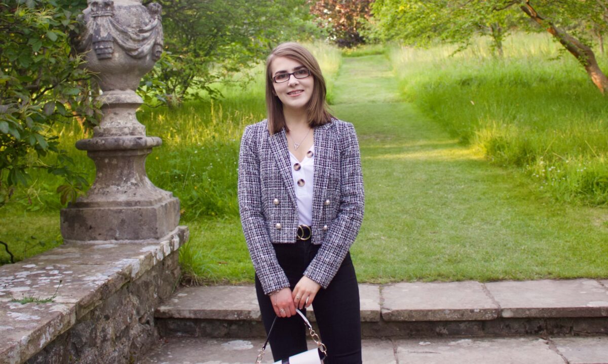 A photo of Elin standing on stone steps which lead to a green patch of land, she is wearing a white v-neck top which has buttons on the neckline, a grey boucle jacket and black skinny jeans. She is smiling at the camera