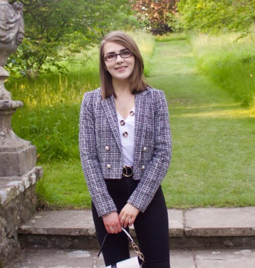 A photo of Elin standing on stone steps which lead to a green patch of land, she is wearing a white v-neck top which has buttons on the neckline, a grey boucle jacket and black skinny jeans. She is smiling at the camera