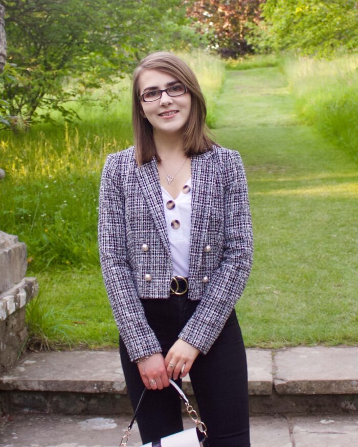 A photo of Elin standing on stone steps which lead to a green patch of land, she is wearing a white v-neck top which has buttons on the neckline, a grey boucle jacket and black skinny jeans. She is smiling at the camera