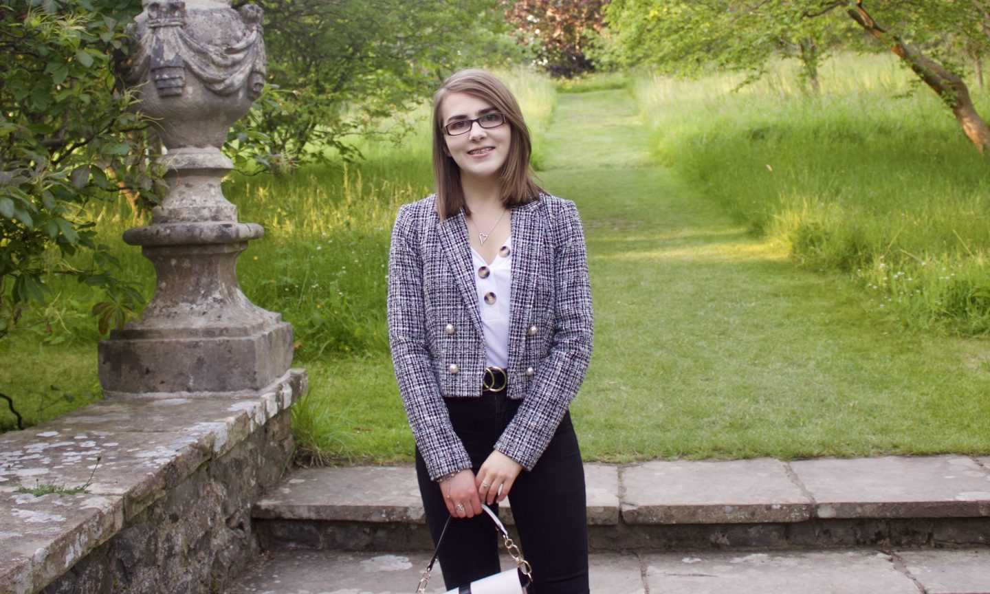 Disability & employment: A photo of Elin standing on stone steps which lead to a green patch of land, she is wearing a white v-neck top which has buttons on the neckline, a grey boucle jacket and black skinny jeans. She is smiling at the camera