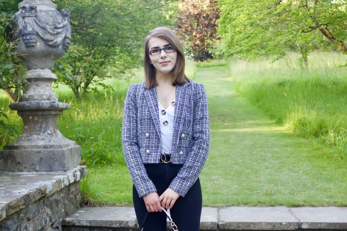 A photo of Elin standing on stone steps which lead to a green patch of land, she is wearing a white v-neck top which has buttons on the neckline, a grey boucle jacket and black skinny jeans
