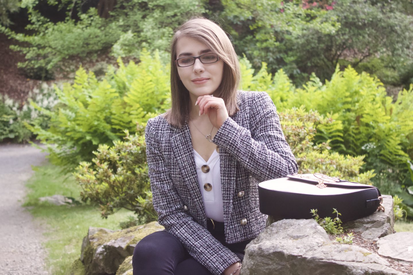 A photo of Elin sitting on a rock and looking down, there are green plants and flowers in the background
