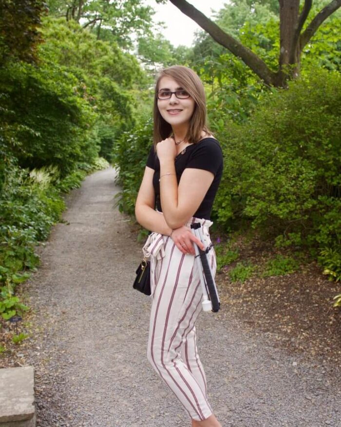 A photo of Elin standing on a path which leads through trees and flowers. She is wearing skinny cream trousers with red stripes, a black bardot top with buttons on the neckline and a black handbag