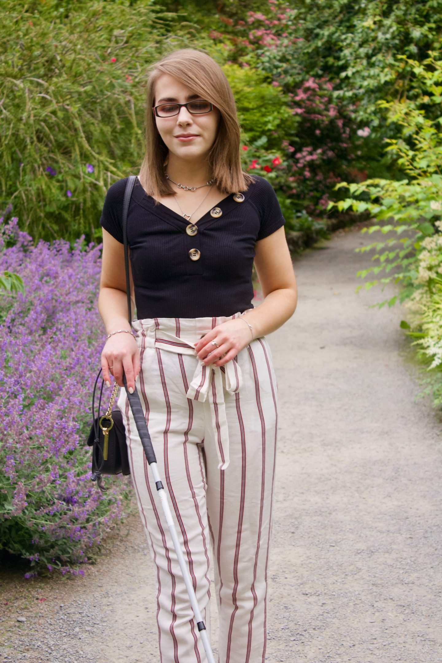 A photo of Elin walking with her cane, she is looking down and is wearing cream trousers with red stripes and a black bardot top which has buttons on the neckline