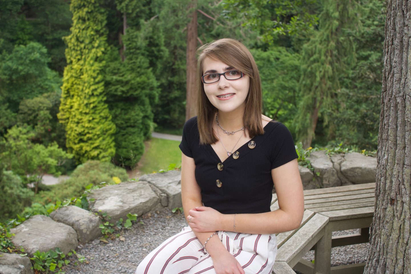 A photo of Elin sitting on a wooden bench which circles around a tree, there are trees in the background